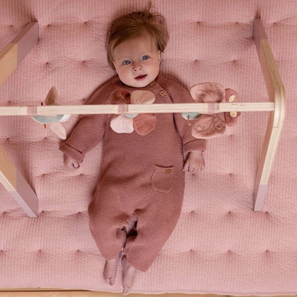 Baby speelt onder houten babygym op roze mat.