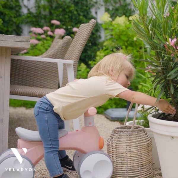 Kind op loopfiets reikt naar plant in tuin.